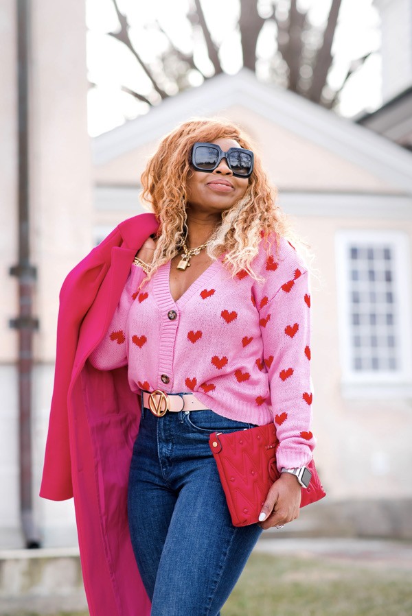 Black woman hearing pink and red hearts sweater with pink Valentino belt and red Valentino clutch and oversized Gucci sunglasses