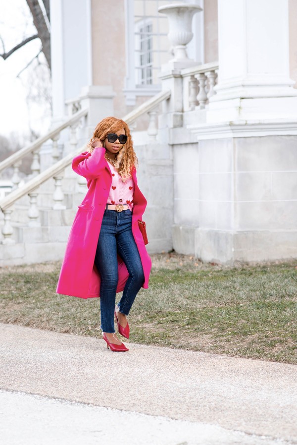 Black woman walking in pink and red hearts sweater and skinny jeans with a pink coat and red pumps